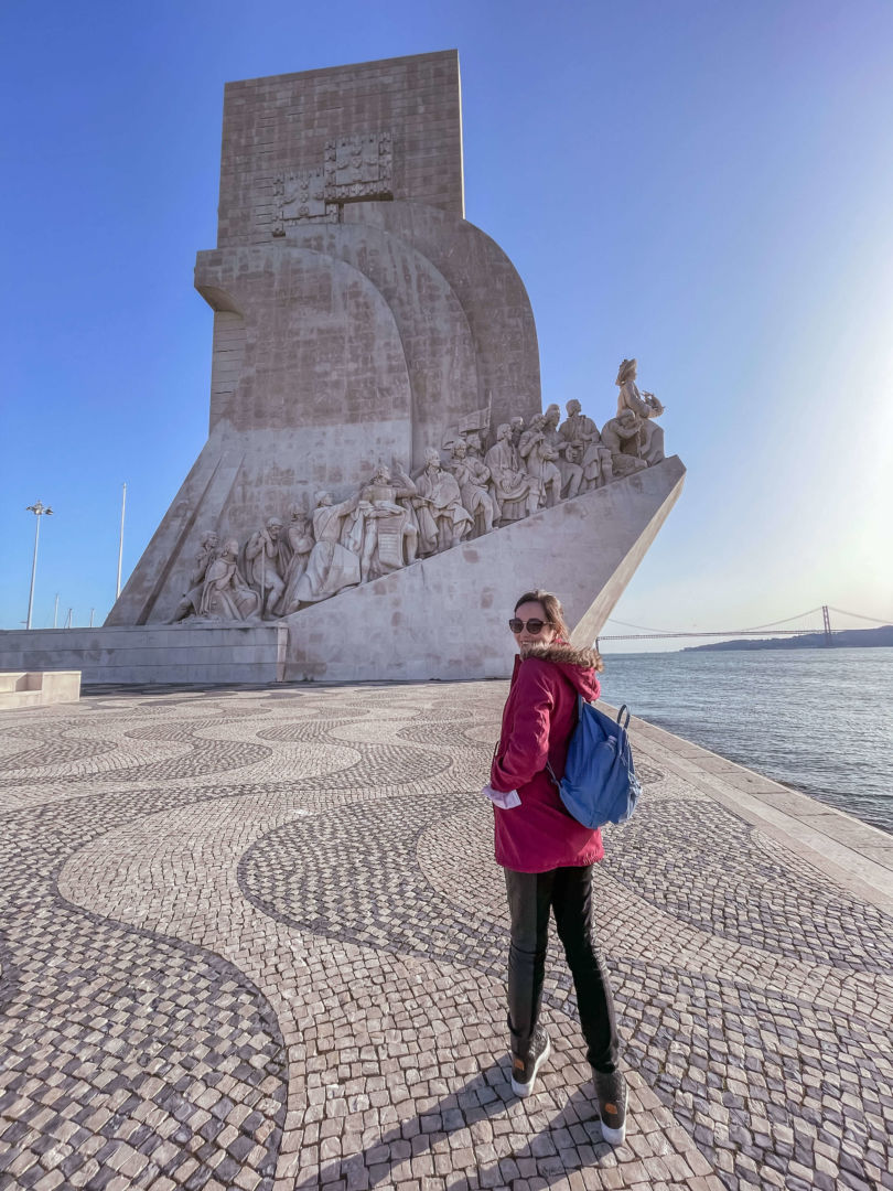 Pessoa de costas e olhando para trás com casaco vermelho e mochila azul. Ao fundo, o monumento do padrão dos descobrimentos e a ponte Vasco da Gama em Lisboa. 
