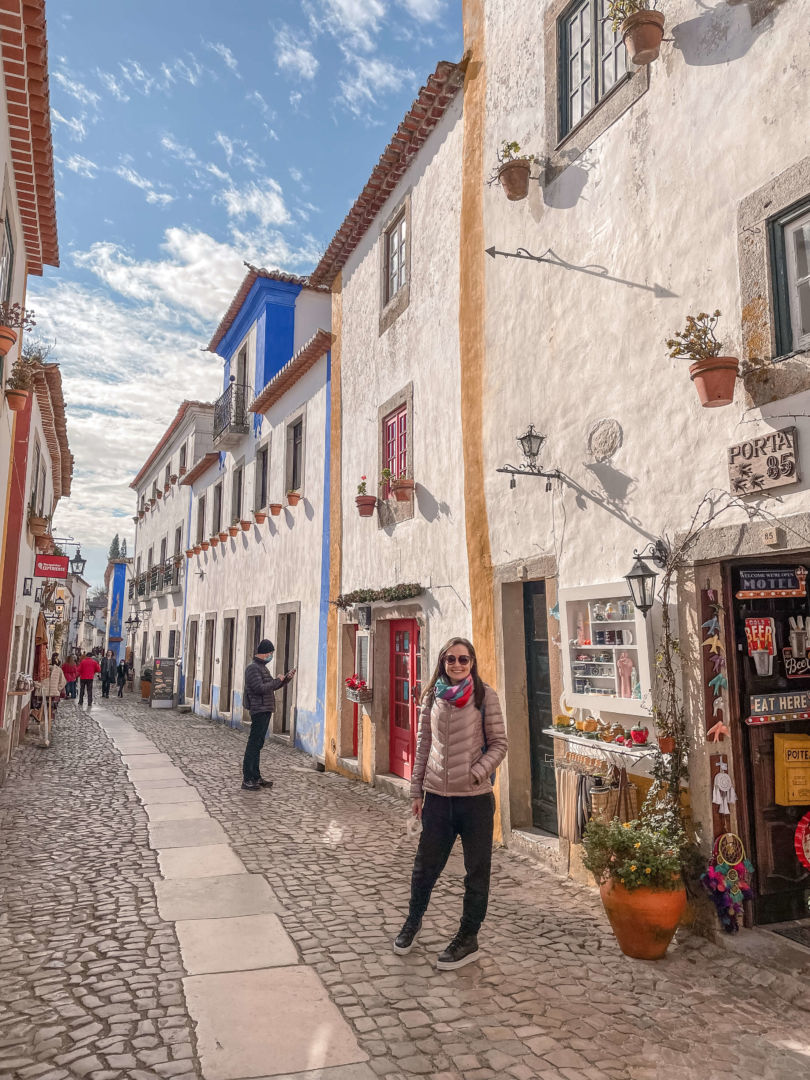 Primeira parada do nosso roteiro de 15 dias de carro entre Portugal e Espanha, em Óbidos. Eu estou em uma rua com construções antigas de dois andares em Óbidos com o céu azul. As construções são brancas com contornos em amarelo e azul. Eu estou posando na frente.