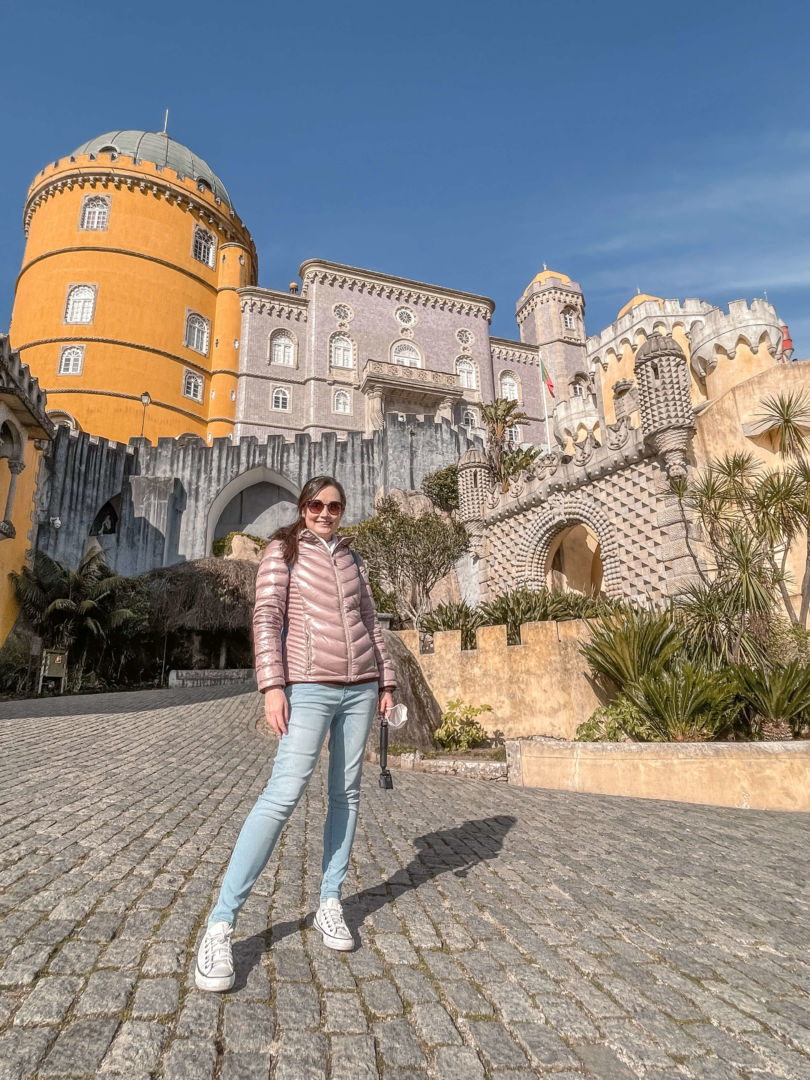 Eu posando em frente ao Palácio da Pena em Sintra. A foto é de baixo, apareço de corpo inteiro e ao fundo o Palácio, com uma cúpula redonda em amarelo e o restante em uma cor entre roxo e Azul. O estilo arquitetônico é o romântico. 