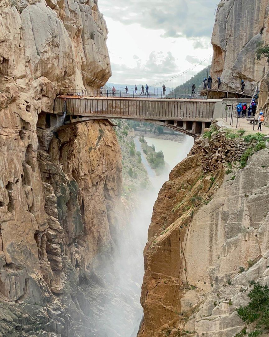 Foto que mostram pessoas atravessando uma ponte ao fundo. A ponte fica em uma altura de dezenas de metros de altura ligando dois desfiladeiros ou rochas. Pessoas passam ao lado, na trilha formada nesse desfiladeiro.