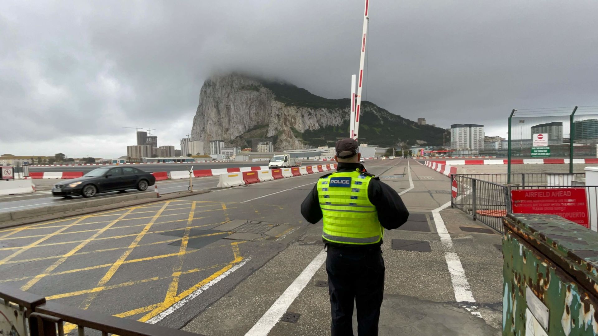 Dia nublado com a pedra de Gibraltar encoberta pela neblina. Em frente, a pista do aeroporto que fica no meio da cidade e um guarda que cuida do trânsito, trancado para a passagem do avião.