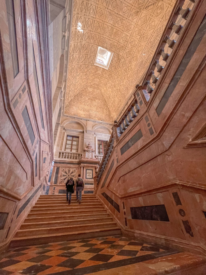 Foto de baixo para cima das escadas do arquivo geral das índias. Uma construção antiga e bonita, com chão de mármore e teto com texturas. 