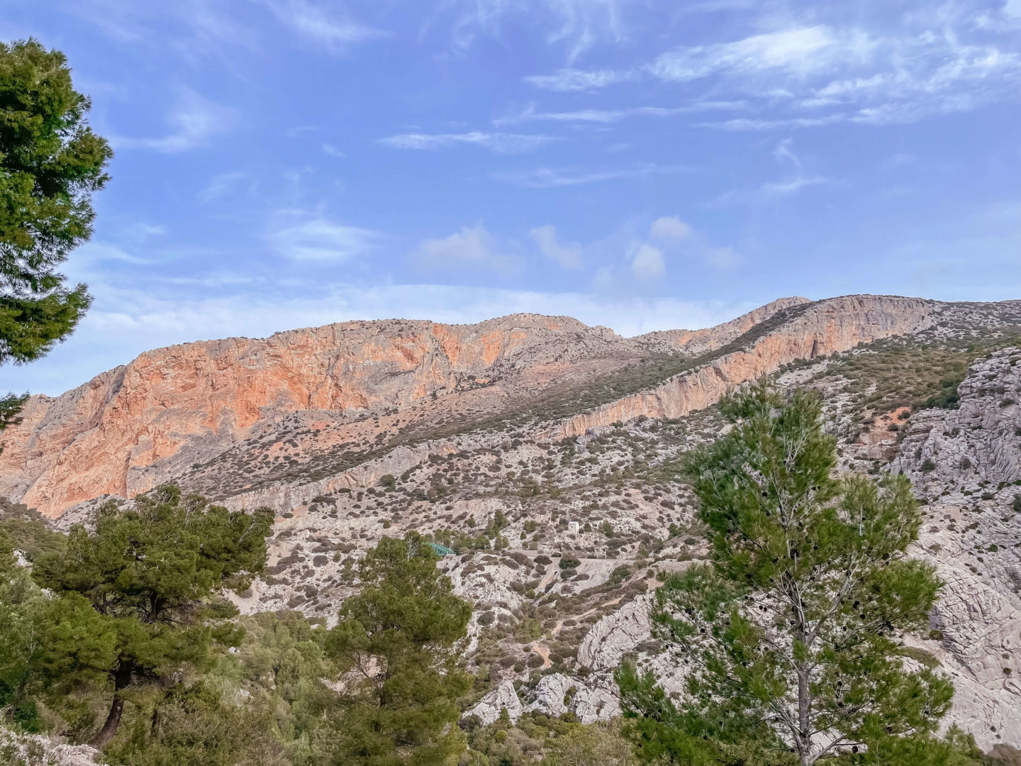 Uma vista bonita paisagem, onde aparece o céu azul, a montanha com tons laranja, cinza e verde e as árvores verdes na frente.