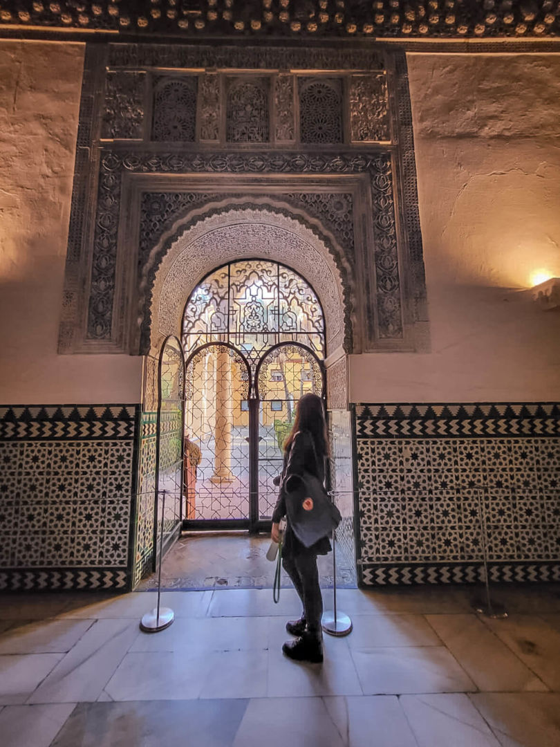 Foto dentro do Real Alcazar. Eu estou de costas em uma janela em arco, onde toda a arquitetura é árabe.
