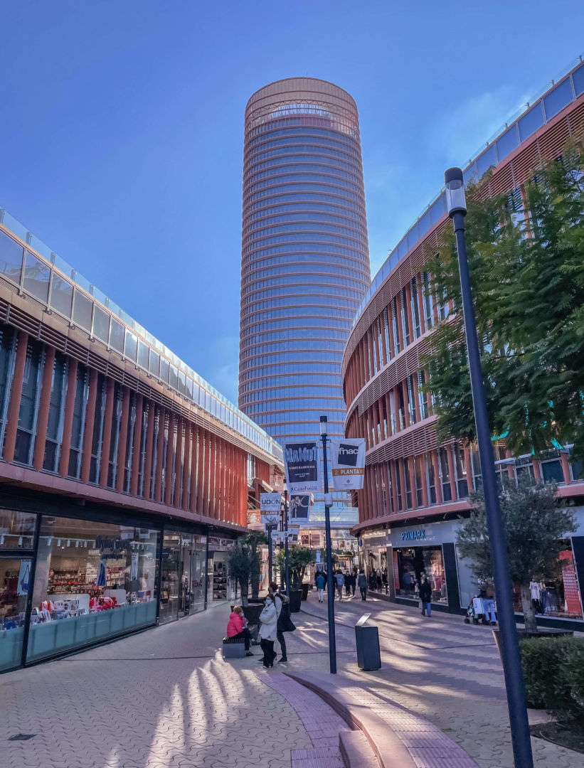 Uma torre no fundo e no centro da foto e um centro comercial a céu aberto com várias lojas nas laterais.