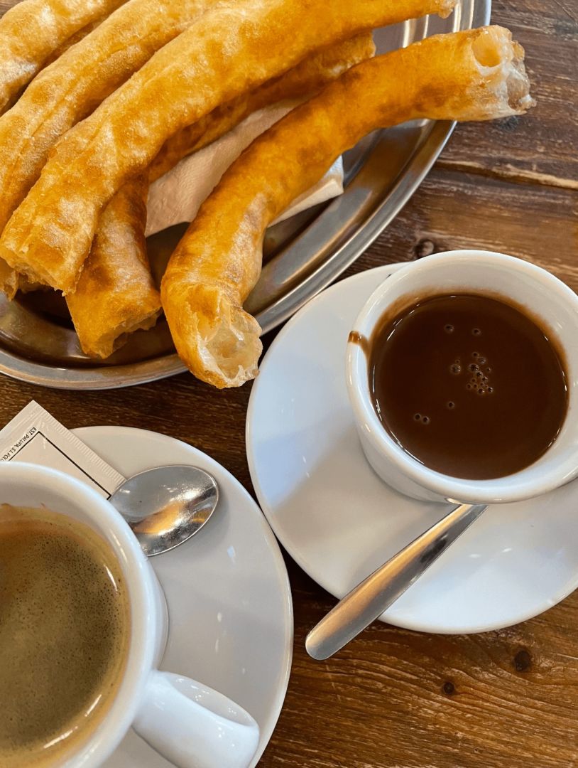 Uma mesa com uma xícara de chocolate quente no centro, uma parte de uma xícara de café e uma parte de um prato de churros.