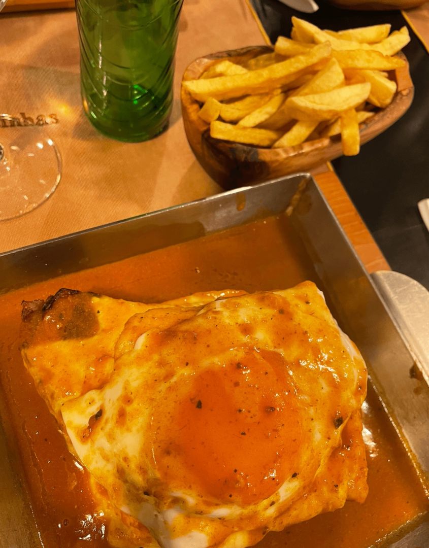 Foto de cima de uma francesinha, o prato típico de Porto. Na imagem é possível ver um prato quadrado com um ovo por cima do sanduíche e muito molho ao redor.