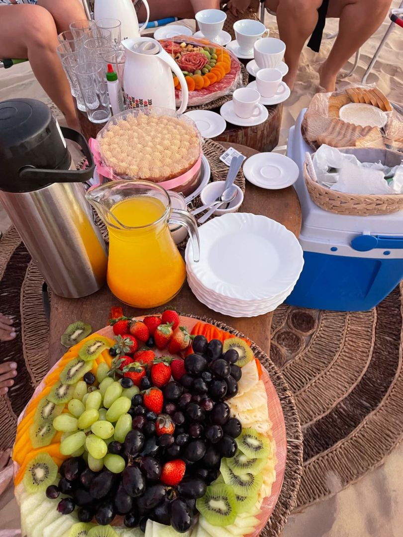 Mesa baixa com uma tábua de frutas com uvas, morangos e kiwis, uma jarra de suco, uma garrafa de café e um bolo decorado. 