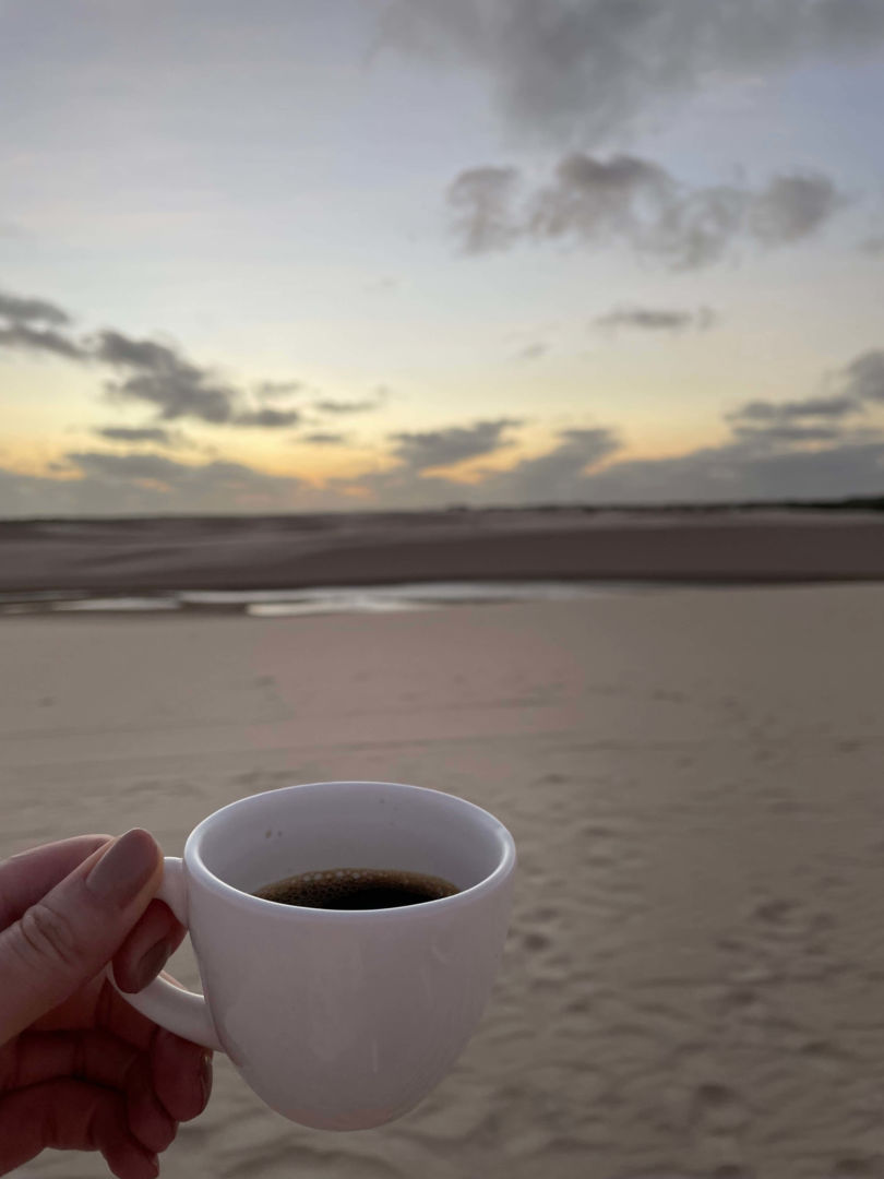 Uma xícara com café preto na frente e os lençóis com o sol nascendo ao fundo.