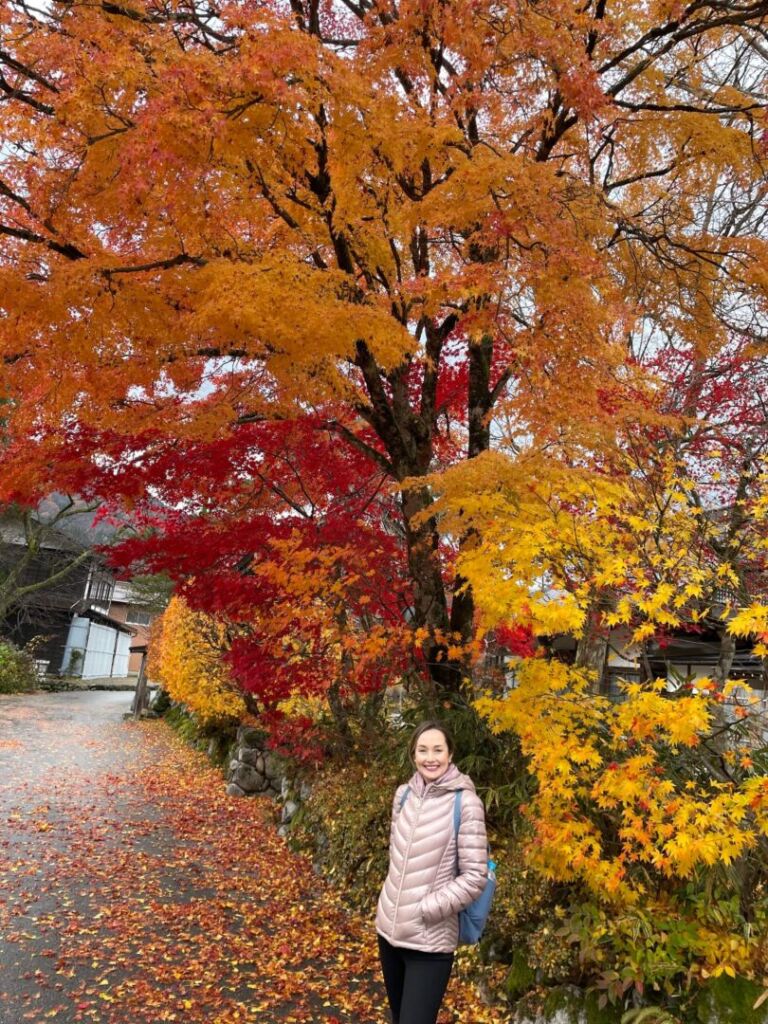 Eu sorrindo na parte inferior da foto em frente a uma árvore grande com as folhas amarelas, laranjas e vermelhas. Tem algumas folhas caídas no chão.