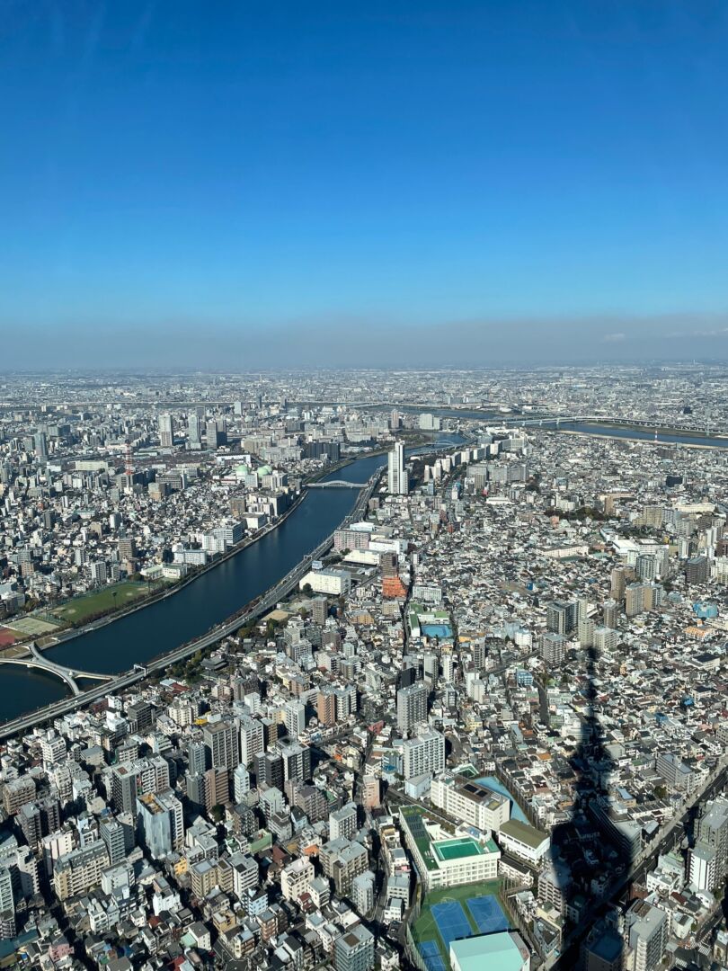 Tóquio vista de cima. A cidade do alto, com um rio passando à esquerda e a sombra da torre Sky Tree no canto direito. O céu está bem azul da metade da foto para cima