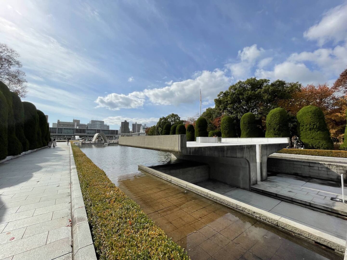 No parque da paz em Hiroshima. O céu está azul com algumas nuvens. Na foto, a chama da paz no canto direito e o museu da paz ao fundo.