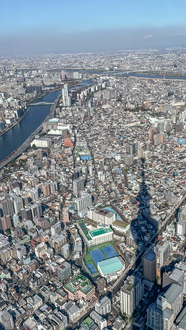 Foto do alto onde se vê Tóquio de cima, céu azul, um rio passando à esquerda e a sombra da Sky Tree. 
