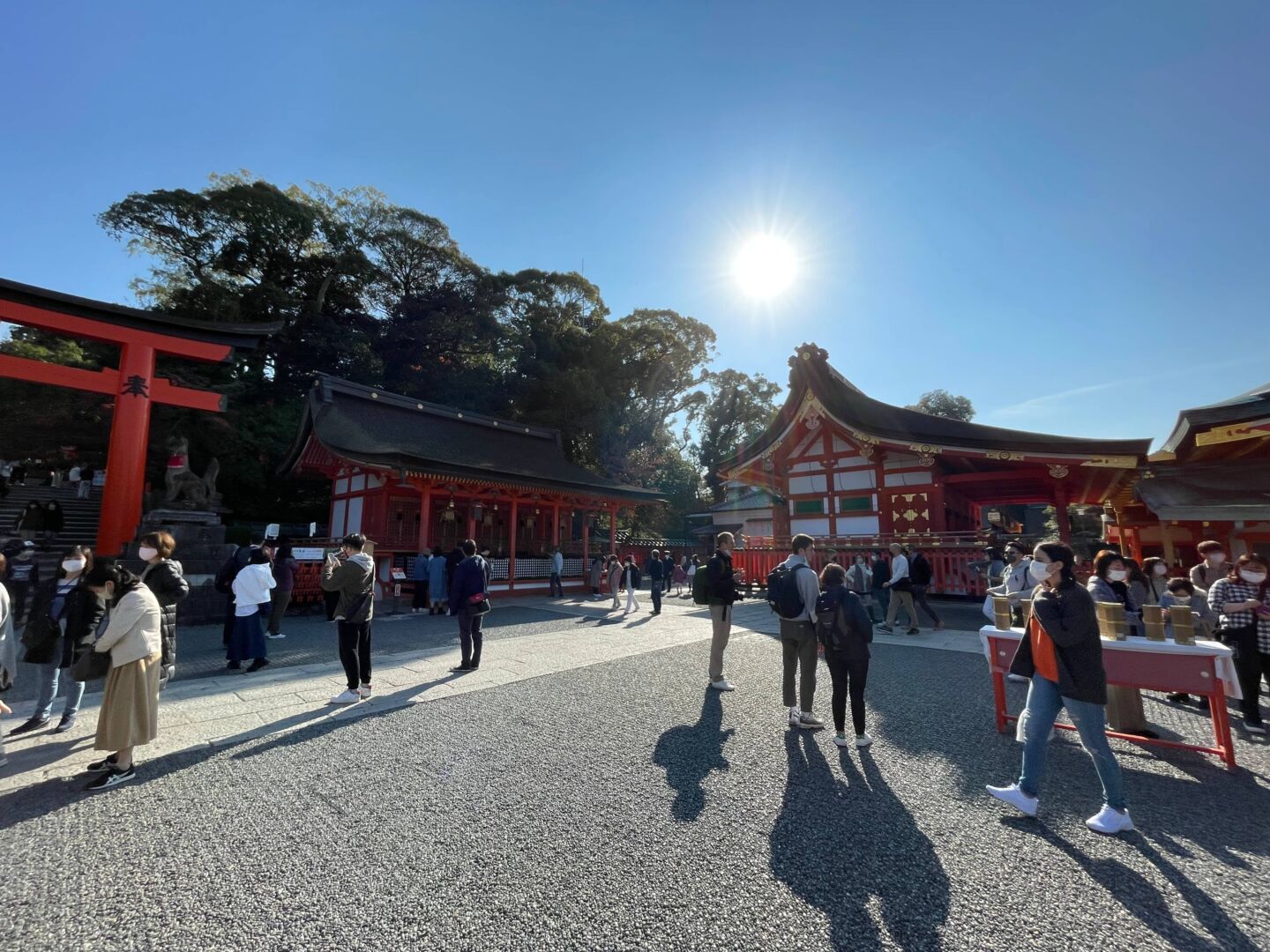Foto contra o sol do templo Inari, tem várias pessoas e todas as construções são vermelho e branco.