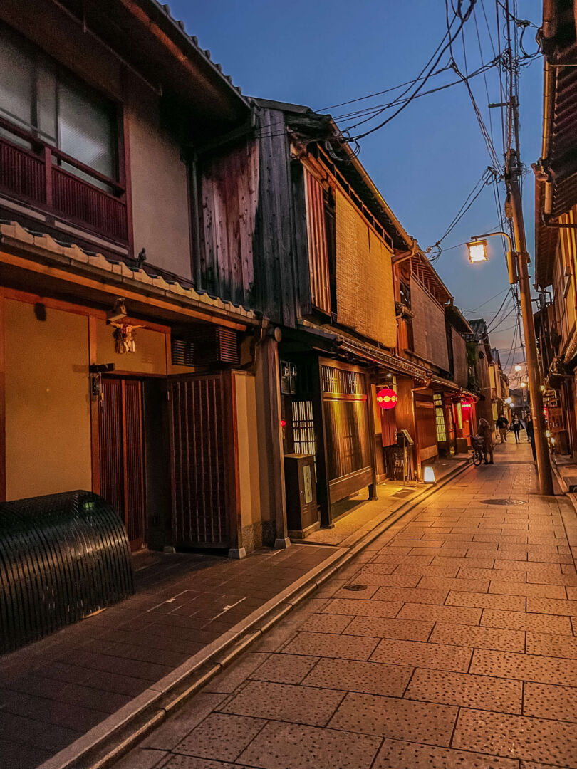 A noite no bairro Gion. A rua é estreita e está vazia. Aparecem casas de dois andares típicas japonesas