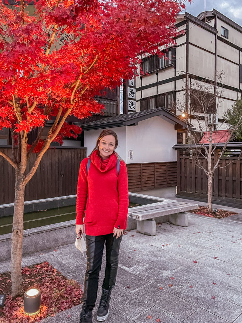 Eu posando para a foto usando uma blusa de lã vermelha ao lado de uma árvore com as folhas no mesmo tom de vermelho.