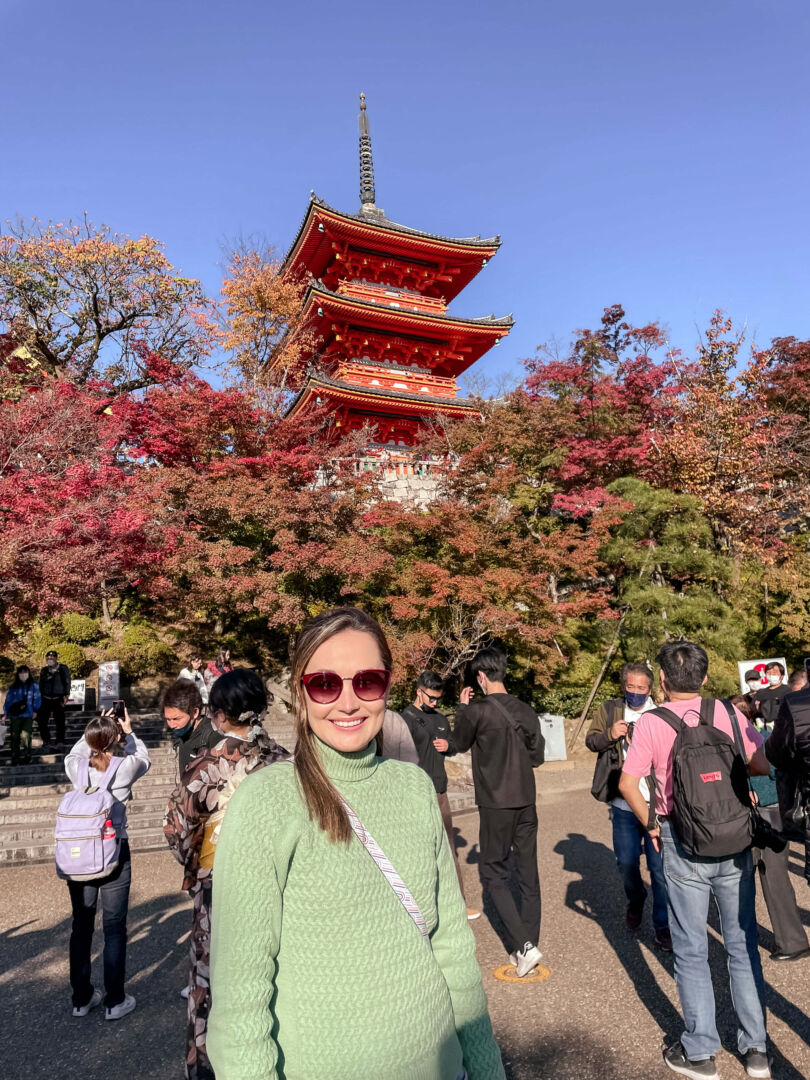 Eu em frente e atrás de mim uma pagoda em vermelho cercada de árvores de folhas vermelhas e verdes.
