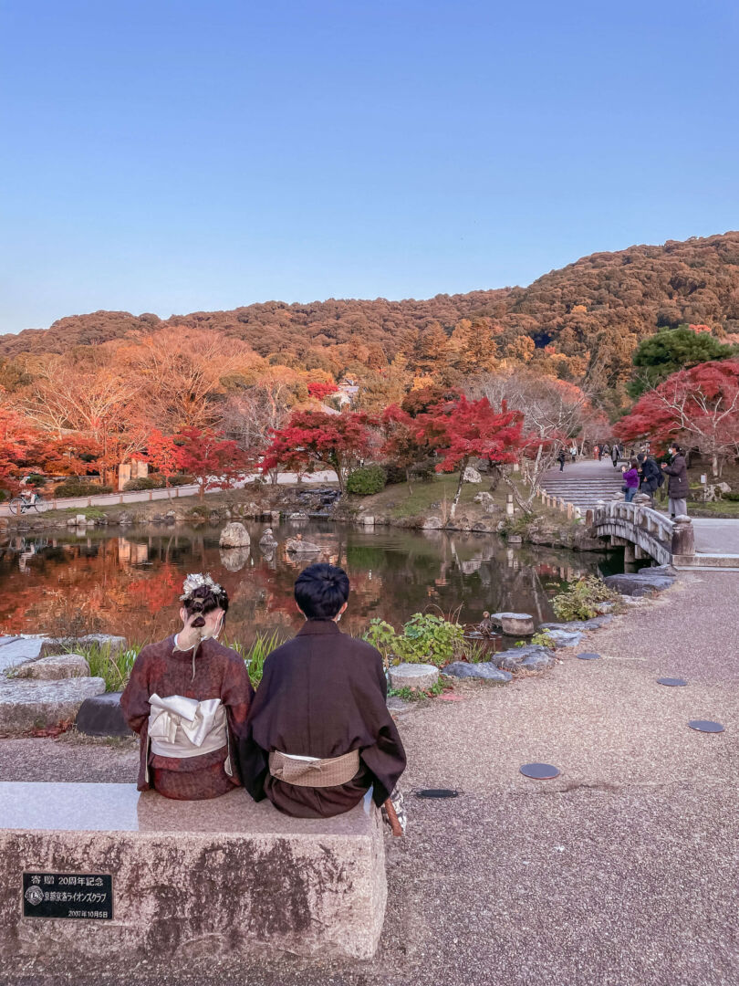 Foto em um parque, com um casal vestido com kimono sentado em um banco em primeiro plano. Ao fundo, um lago, uma pequena ponte e árvores de todas as cores.