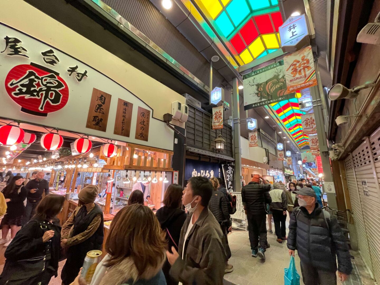 Foto interna do Nishiki Market, com o teto em vidro colorido e uma barraquinha de comida em primeiro plano com pessoas passando na frente.