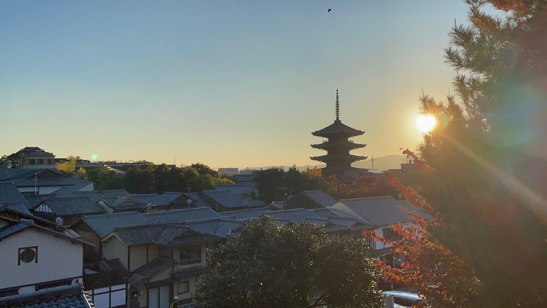 foto no pôr do sol onde se vê o contorno da cidade, com o sol aparecendo entre as àrvores no canto direito e o contorno de uma pagoda ao fundo