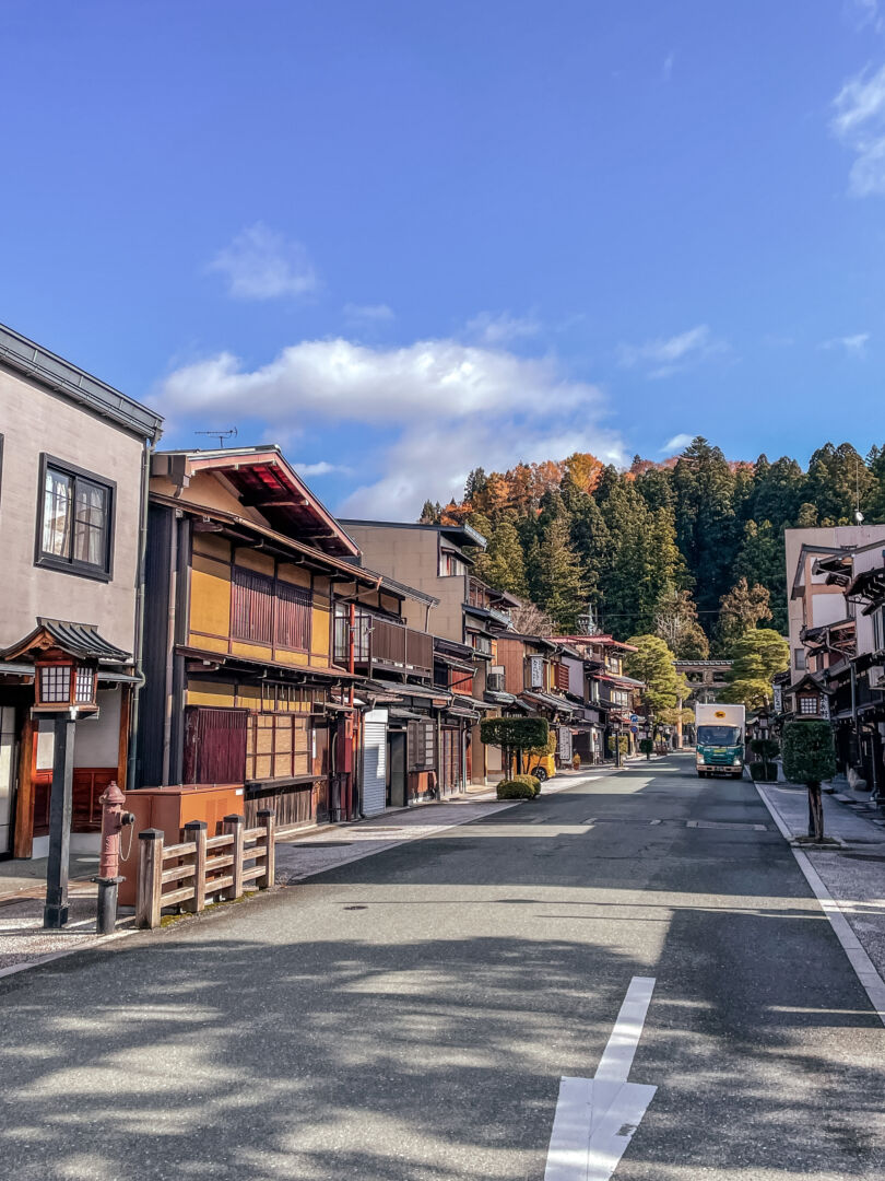 Foto em uma rua de Takayama, com casas históricas e o céu azul e árvores verdes e com folhas amarelas ao fundo.