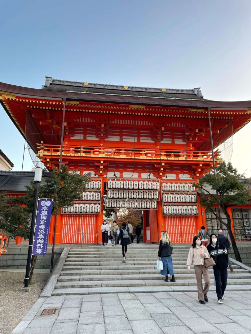 O templo Yakata bem ao centro, de cor vermelha e com várias lanternas japonesas em frente. É um final de tarde e o céu está azul claro. Em frente uma pequena escada com pessoas descendo e subindo.