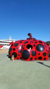 escultura de uma abobora gigante vermelha com pontos pretos, criança brincando, ferry boat ao fundo