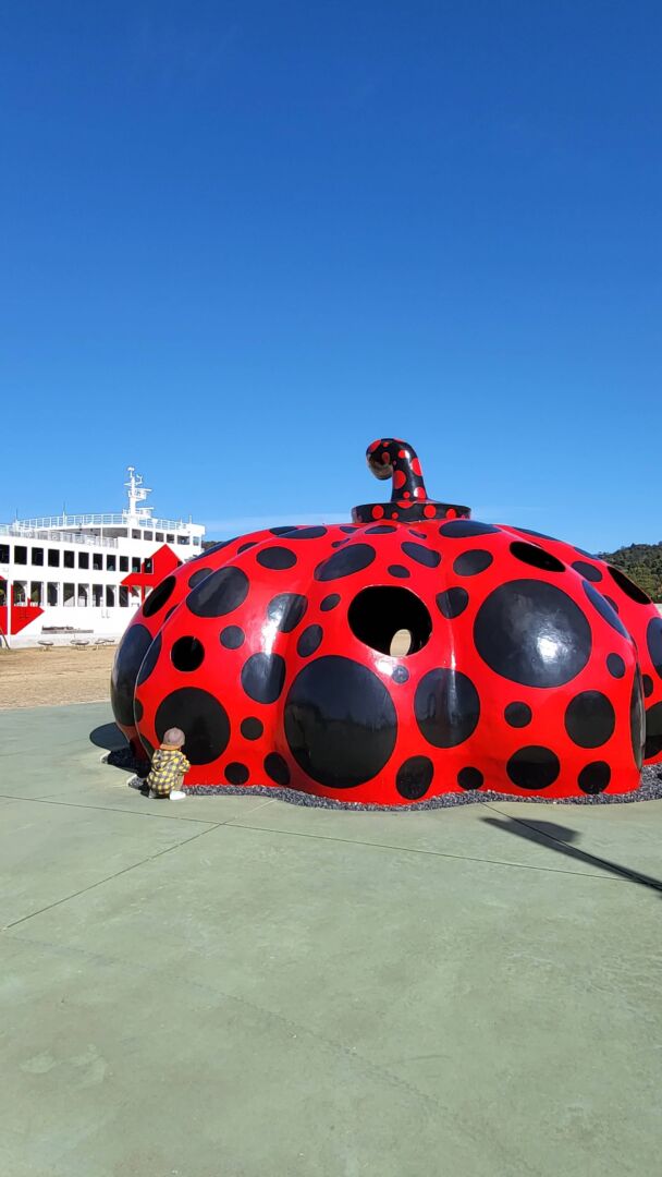 escultura de uma abobora gigante vermelha com pontos pretos, criança brincando, ferry boat ao fundo