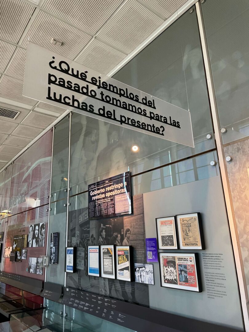 Por dentro do Museu, onde se lê uma placa escrita: "Qué ejemplos del pasado tomamos para las luchas del presente?"com alguns quadros do museu ao fundo.