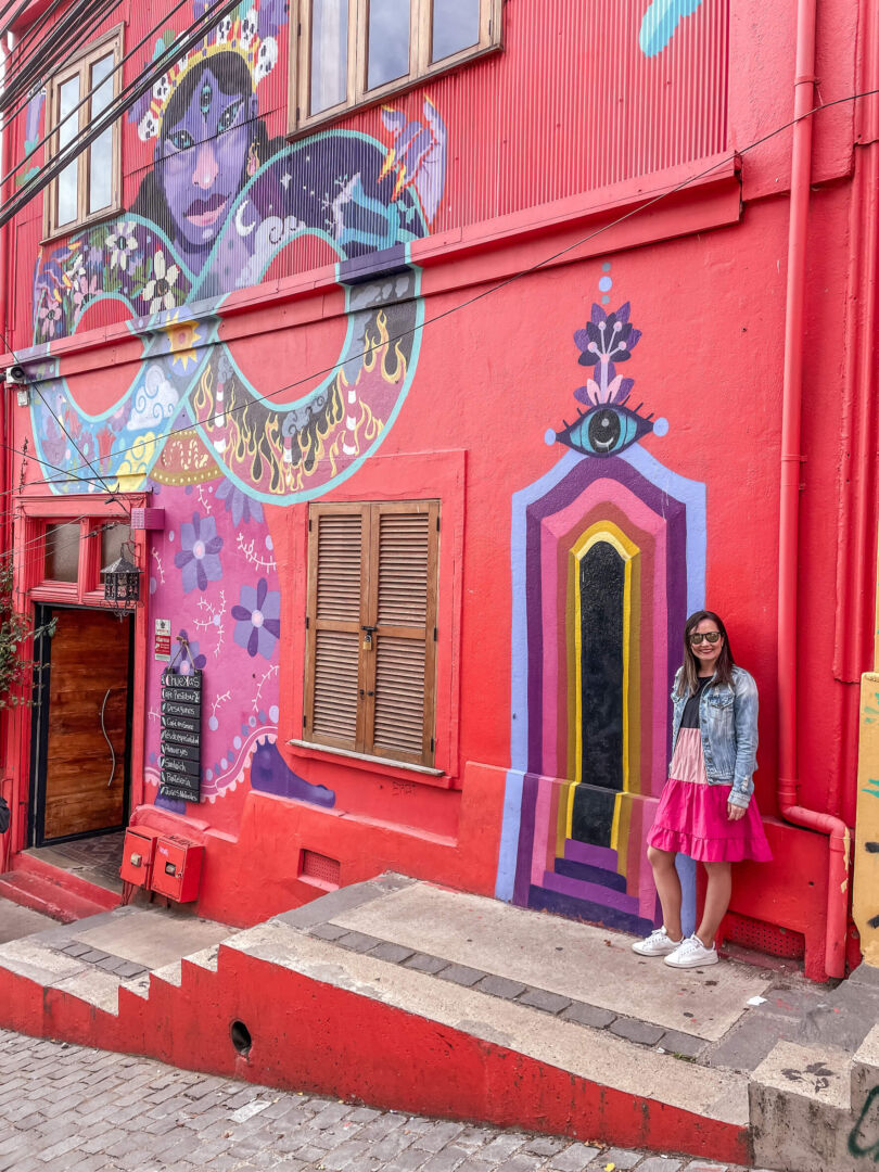 Eu em frente a uma fachada colorida em Valparaíso. é uma grande casa vermelha, com um desenho mais colorido simulando uma porta.